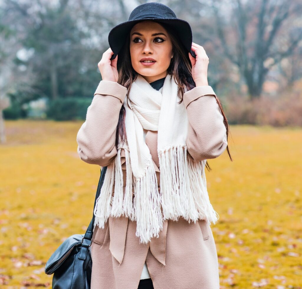 "A woman wearing a camel coat, white scarf, black hat, and a black shoulder bag standing in a park with autumn leaves on the ground."