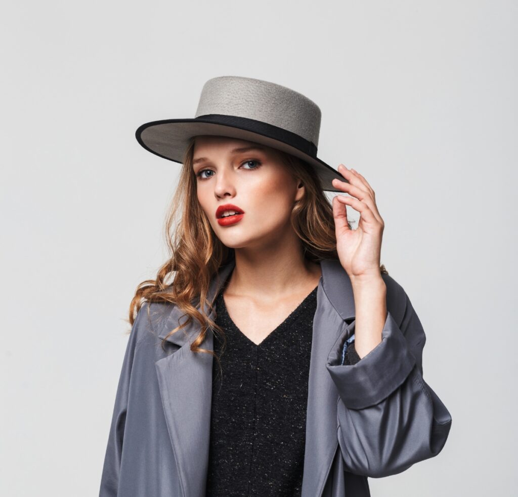 A stylish woman wearing a wide-brim gray gambler hat with a black band, paired with a loose gray trench and a black speckled top.