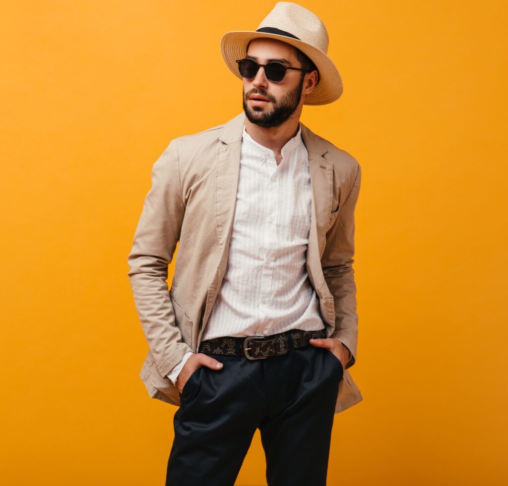 A stylish man posing against a solid orange background. He wears a light beige blazer, white linen shirt, dark trousers, a braided belt, a straw hat, and dark sunglasses, with hands in his pockets.