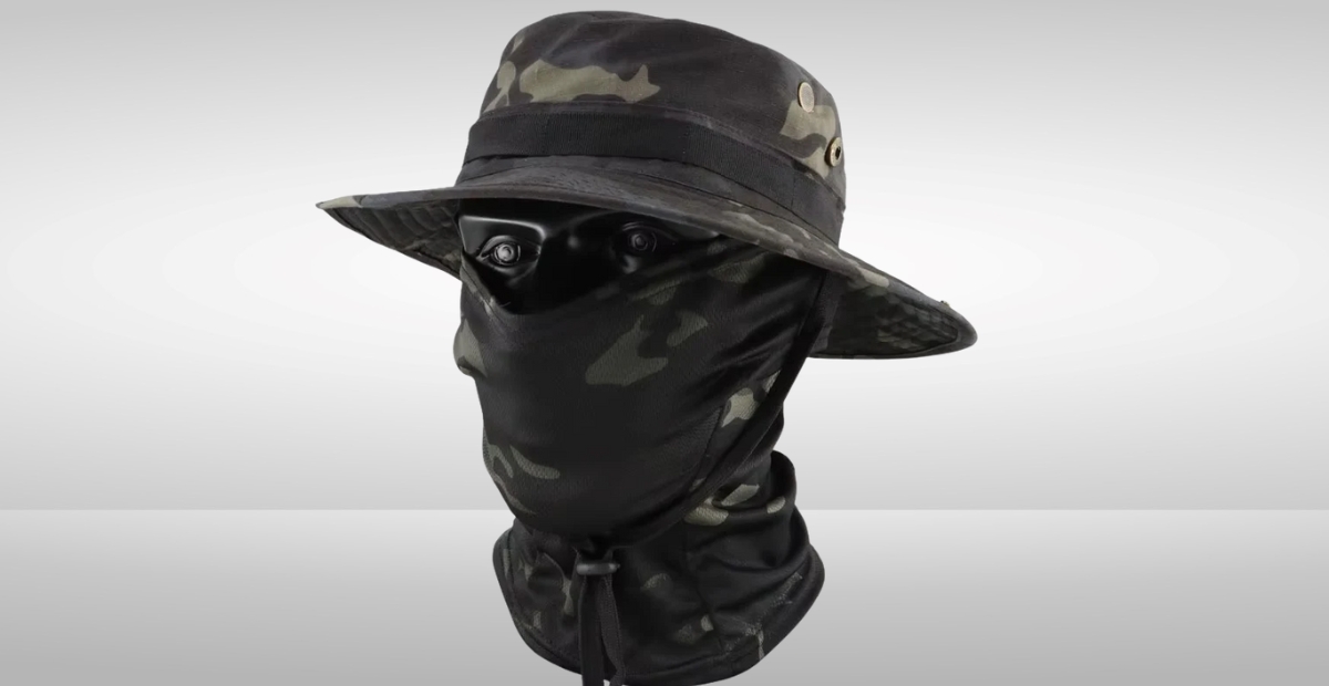 Eine Kombination aus einer getarnten Sturmhaube und einer Mütze. Die Sturmhaube bedeckt das Gesicht, sodass nur die Augen sichtbar sind, und die Mütze im Bucket-Stil mit Camouflage-Muster sitzt auf dem Kopf.