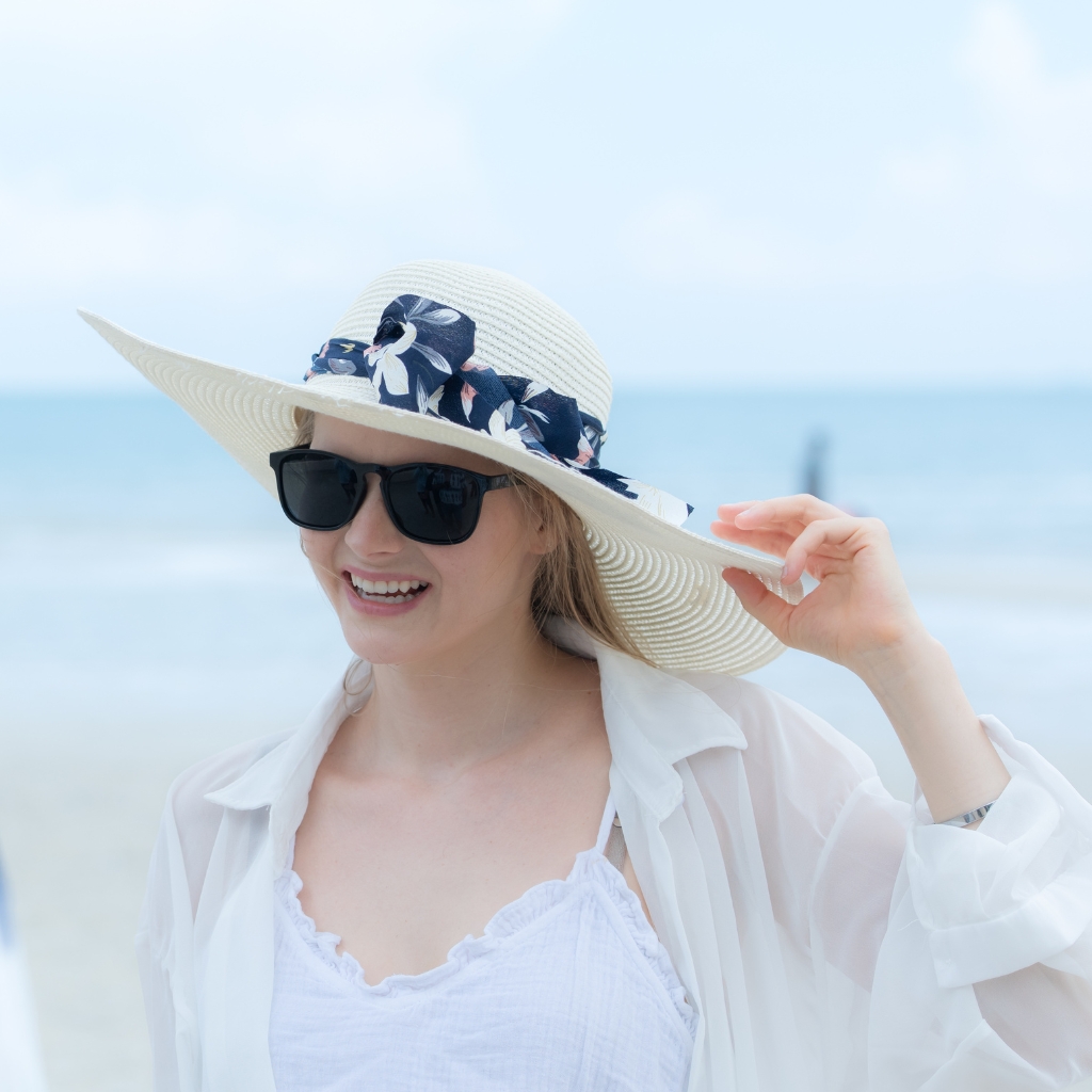 A smiling woman wearing a wide-brimmed straw hat with a dark blue floral ribbon, black sunglasses, and a light white cover-up on a beach backdrop with the sea in the distance.