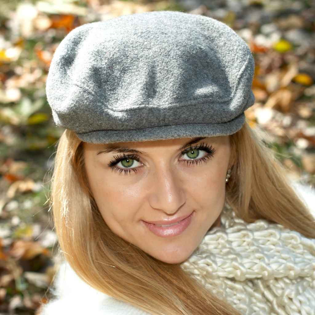 Une femme souriante aux yeux verts, coiffée d'une casquette grise et portant une écharpe tricotée couleur crème, photographiée en plein air sous un soleil radieux, avec des feuilles d'automne en arrière-plan.

