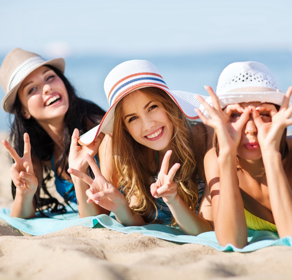 Three smiling women lying on a beach blanket wearing wide-brim sun hats; blue sea in the background.