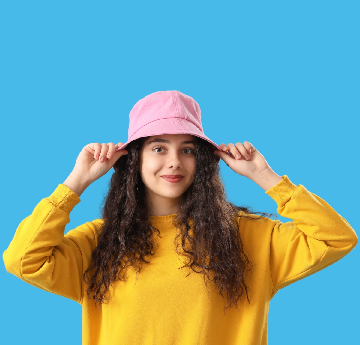 “Young woman wearing a yellow sweatshirt and pink bucket hat, posing against a bright blue background.”