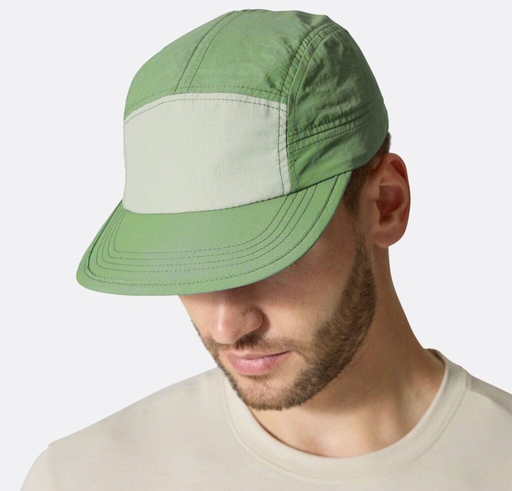 A stack of five-panel caps arranged in a row, each cap made of color-blocked fabric panels in muted tones (blue, beige, gray, brown, green). The caps feature a mid-front color patch and curved brims, with visible stitching and side eyelets.
