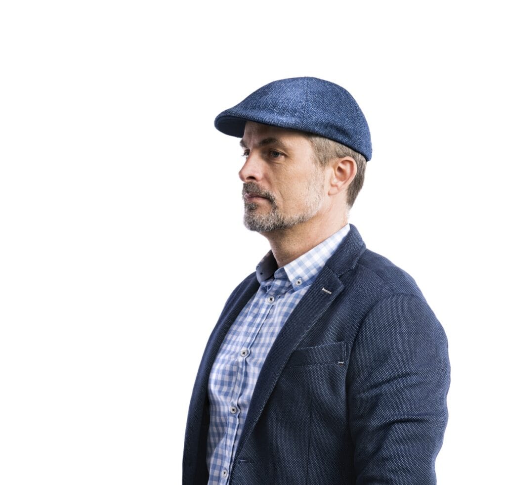 "Side profile of a middle-aged man wearing a blue checkered button-down shirt, a navy textured blazer, and a blue flat cap made by a custom cap manufacturer."