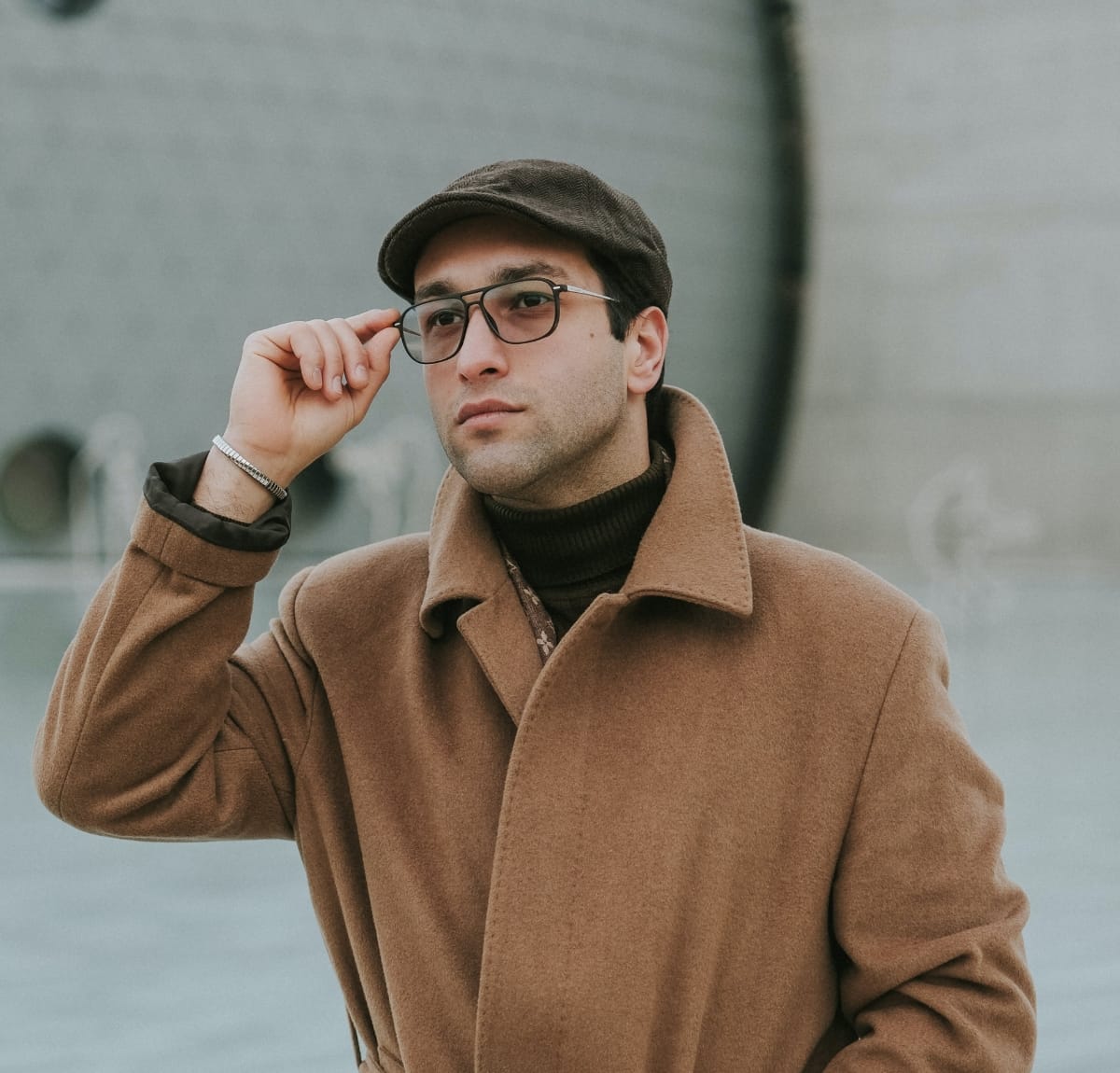 A man wearing a brown wool coat, a dark brown turtleneck, a flat cap, and glasses. He is adjusting his glasses with one hand in an outdoor urban setting.