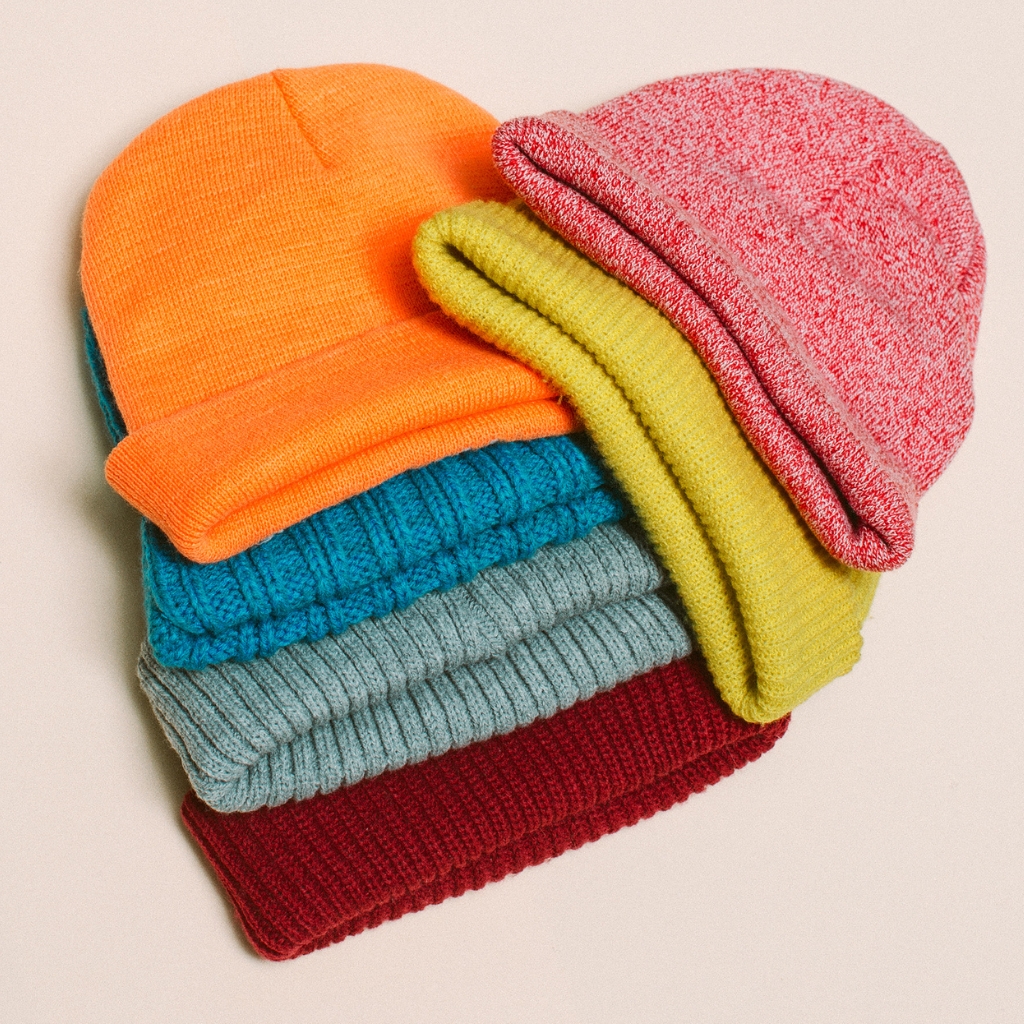 A colorful stack of knit beanies in bright orange, teal, gray, burgundy, with two folded tops forming a casual pile.