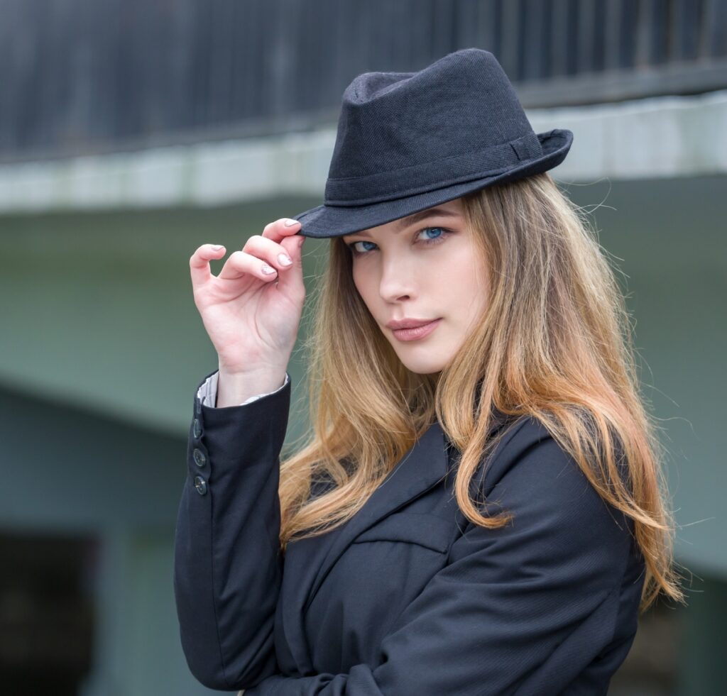 A stylish woman wearing a dark blazer and a custom homburg hat, tipping the hat with her right hand.