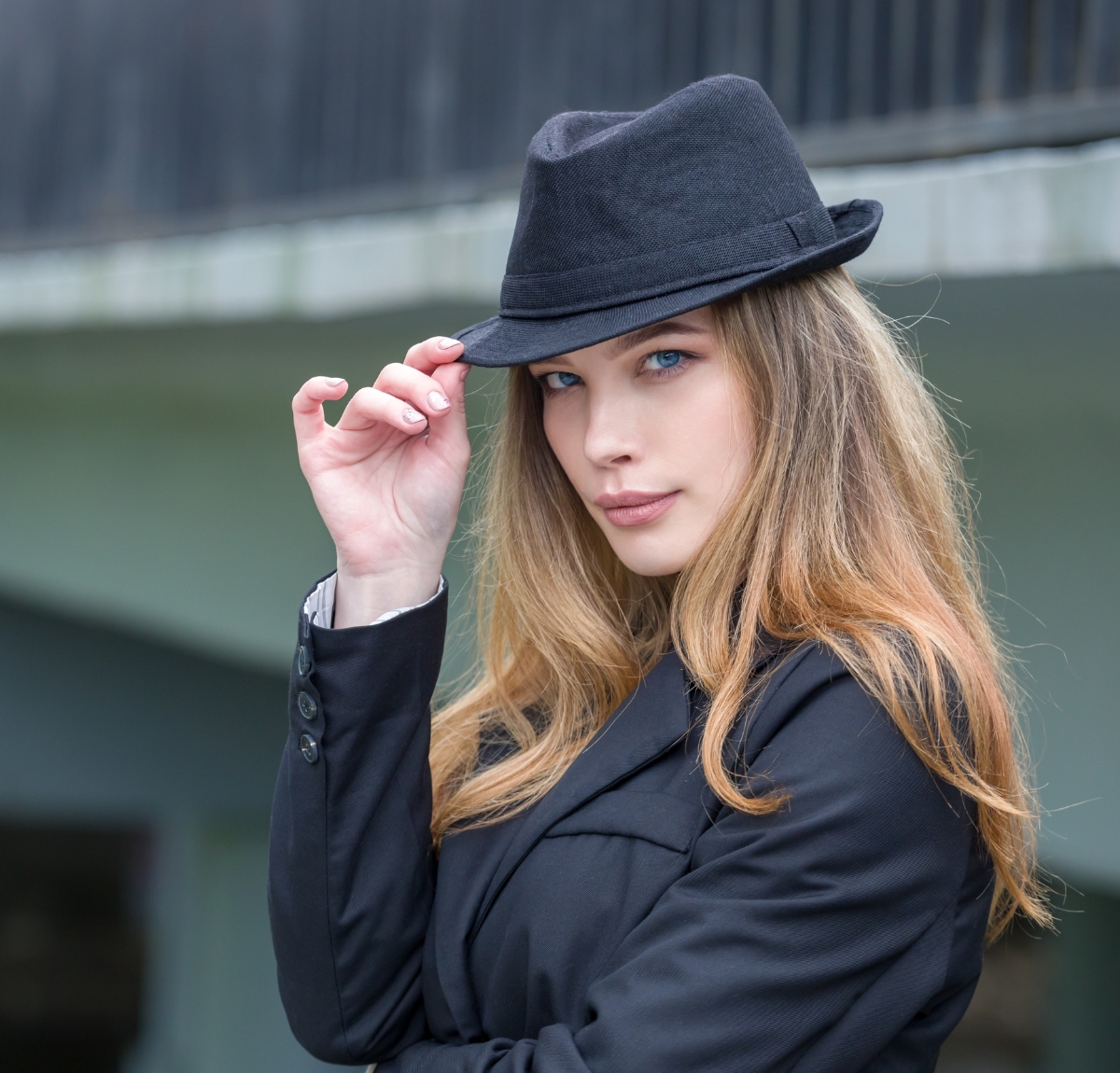 A stylish woman wearing a dark blazer and a custom homburg hat, tipping the hat with her right hand.