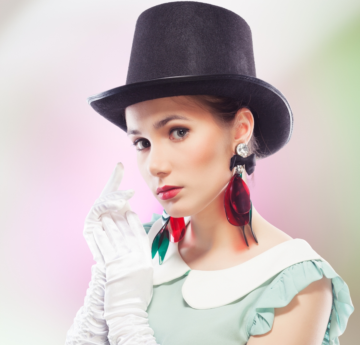 A glamorous woman wearing a tall black top hat, white elbow-length gloves, and bold red earrings. She is looking at the camera with a soft expression against a pastel, blurred background.