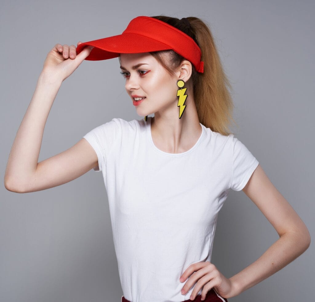 A stylish woman wearing a bright orange visor cap, white t-shirt, and bold yellow lightning bolt earrings, posing with one hand on her hip.