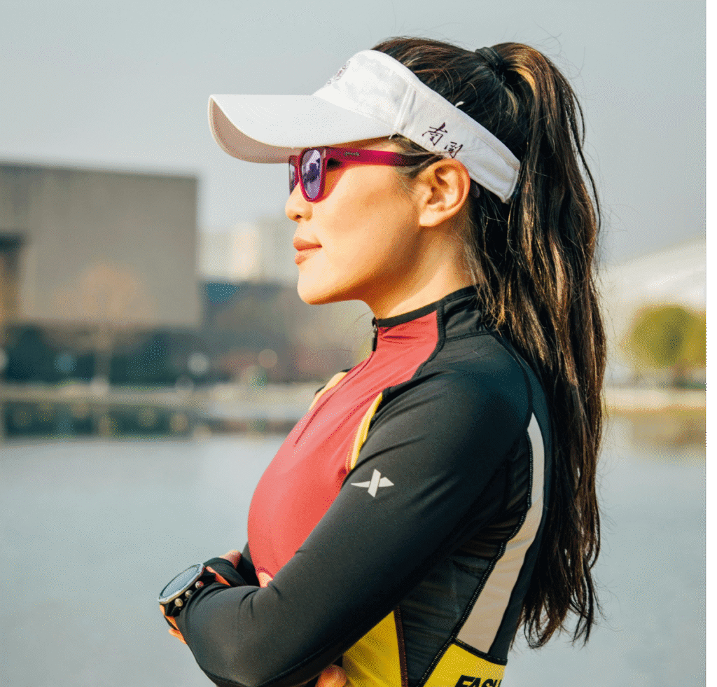 Side profile of a woman outdoors wearing a white visored cap, pink sunglasses, and a color-block athletic outfit. Her arms are crossed in front of her, and a river and modern buildings are blurred in the background.