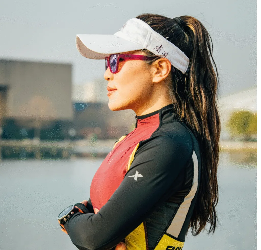 Side profile of a woman outdoors wearing a white visored cap, pink sunglasses, and a color-block athletic outfit. Her arms are crossed in front of her, and a river and modern buildings are blurred in the background.