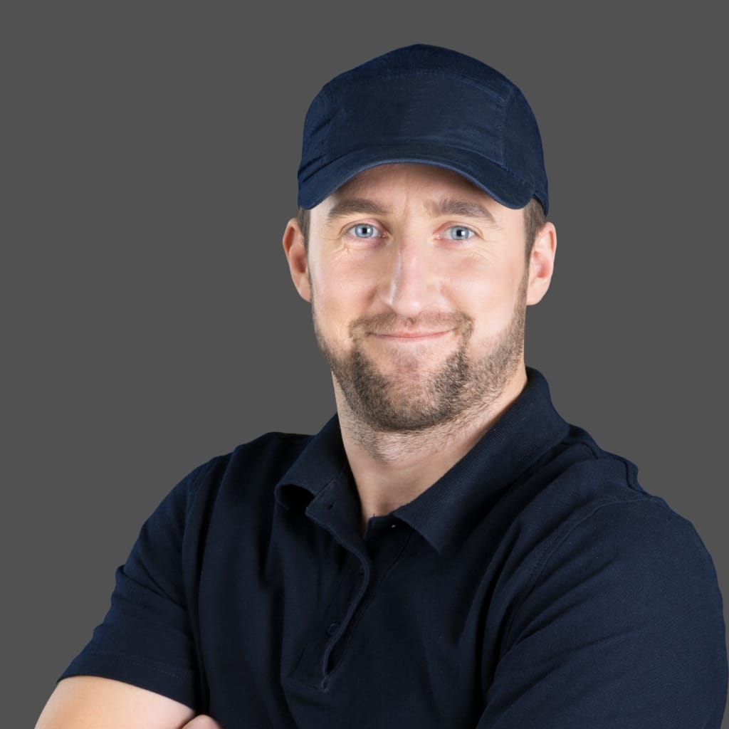 A smiling man wearing a navy dad cap and a dark polo shirt, posing against a neutral gray background.