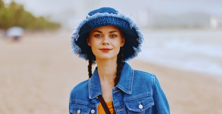Eine Frau an einem Sandstrand, die einen ausgefransten Jeanshut und eine dazu passende Jeansjacke trägt, mit ruhigem Gesichtsausdruck und geflochtenen Haaren.