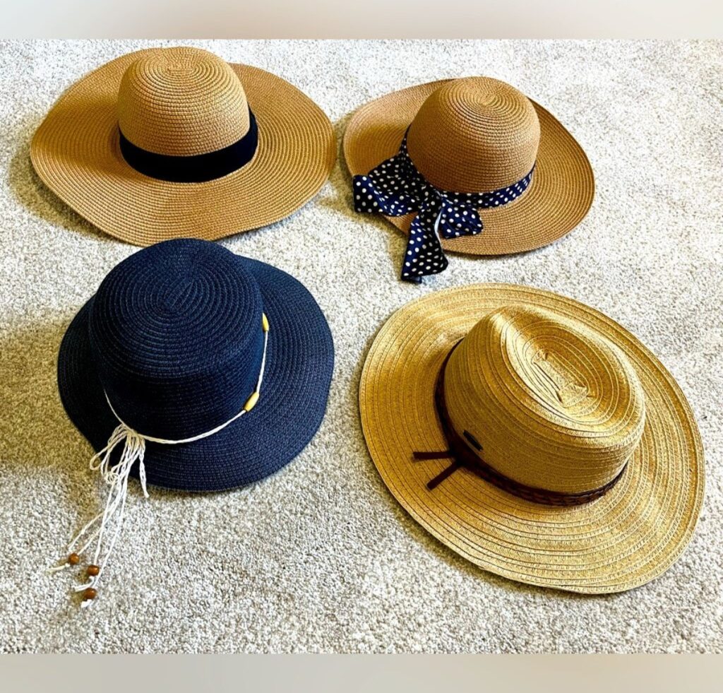 A collection of four floppy-brim straw hats laid on a light carpet: two natural straw hats with simple ribbons, one navy-blue hat with a white decorative cord, and one tan hat with a darker brown band.