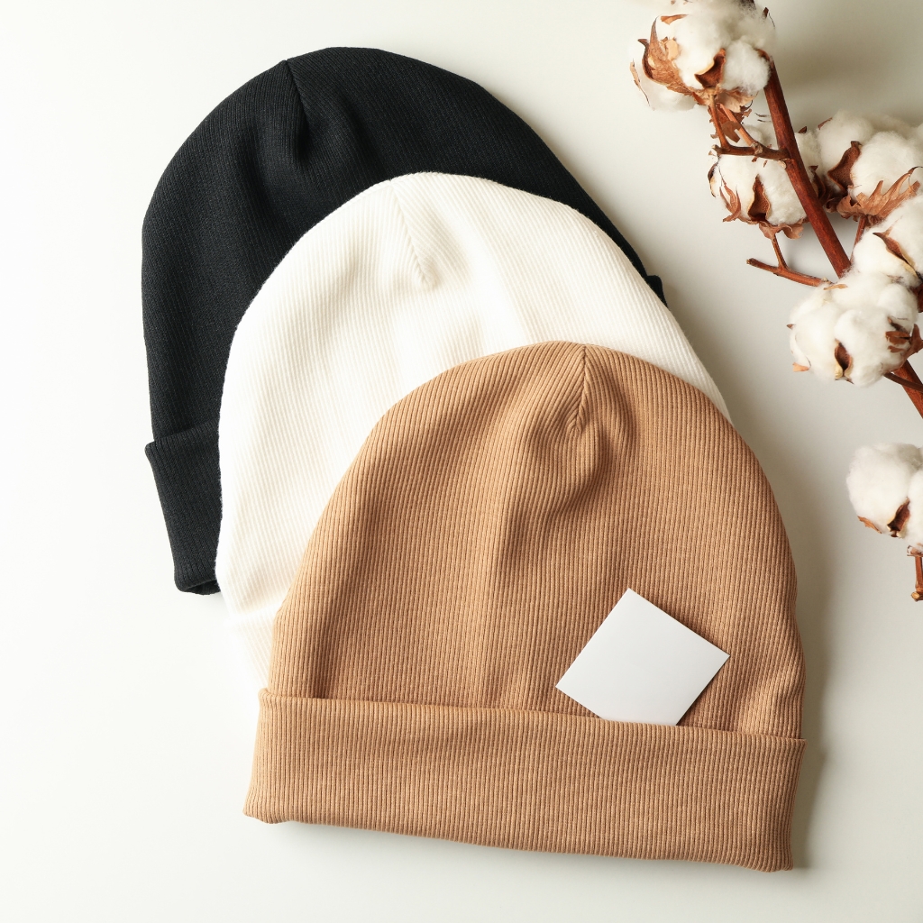 Three ribbed knit fisherman beanies in black, white, and camel colors stacked together, with a small white card tucked into the camel hat and cotton stems in the background.