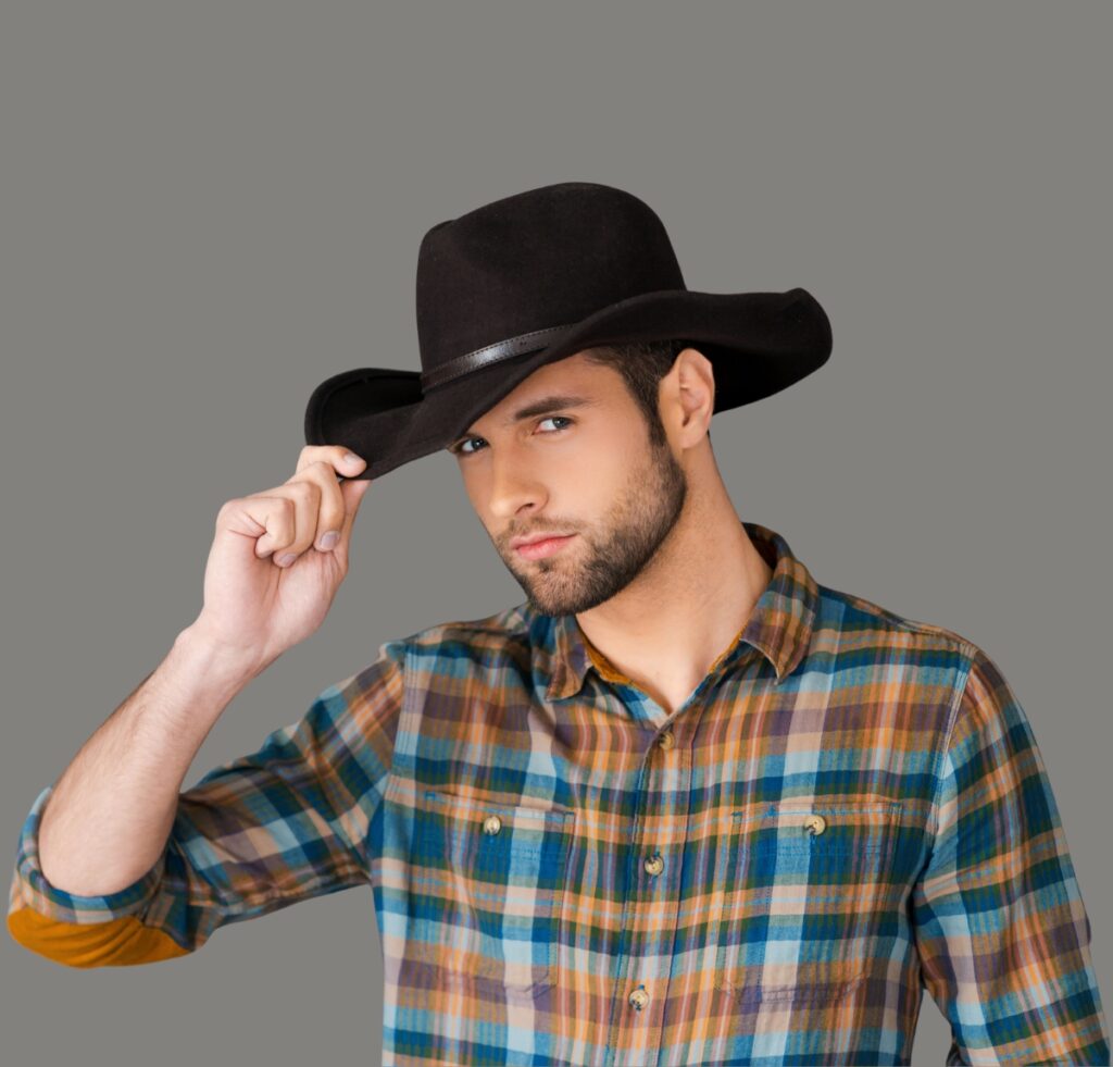 A man wearing a black cowboy hat and a colorful plaid shirt, tipping his hat against a gray background.