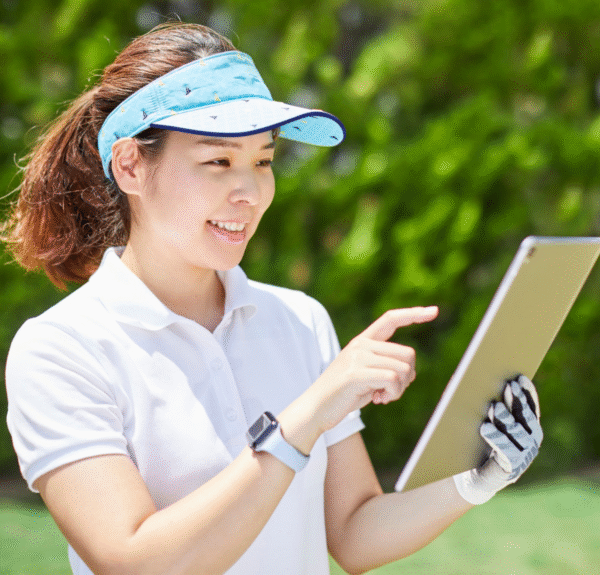 A smiling woman wearing a light blue visor and white polo shirt, outdoors on a sunny day, holding and interacting with a tablet.