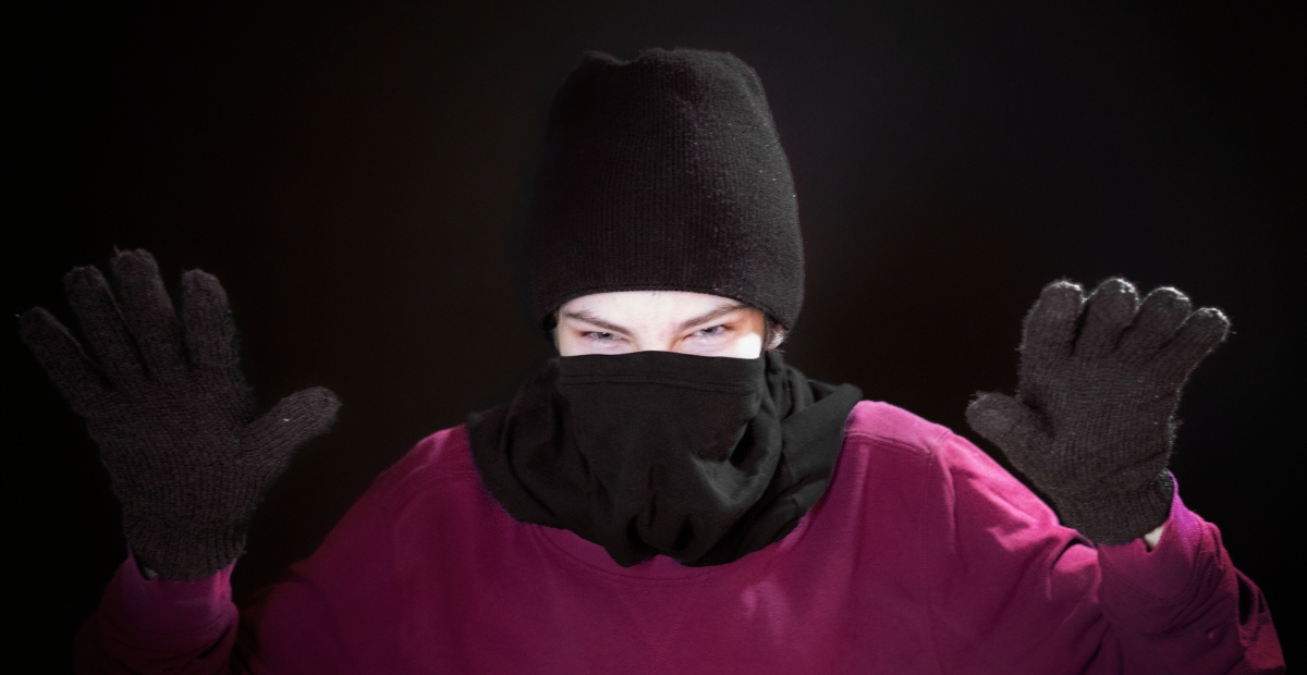Eine Person, die eine schwarze Sturmhaube, ein lila Hemd, und schwarze Strickhandschuhe trägt, mit Händen erhoben vor einem dunklen Hintergrund