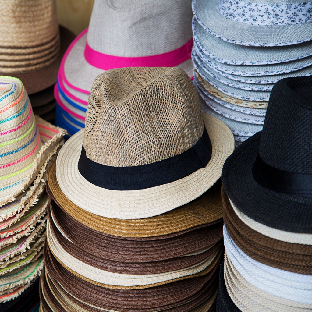 A stack of straw beach hats in natural beige, brown, and black tones, with a central hat featuring a black ribbon band; other hats and woven patterns appear in the background.