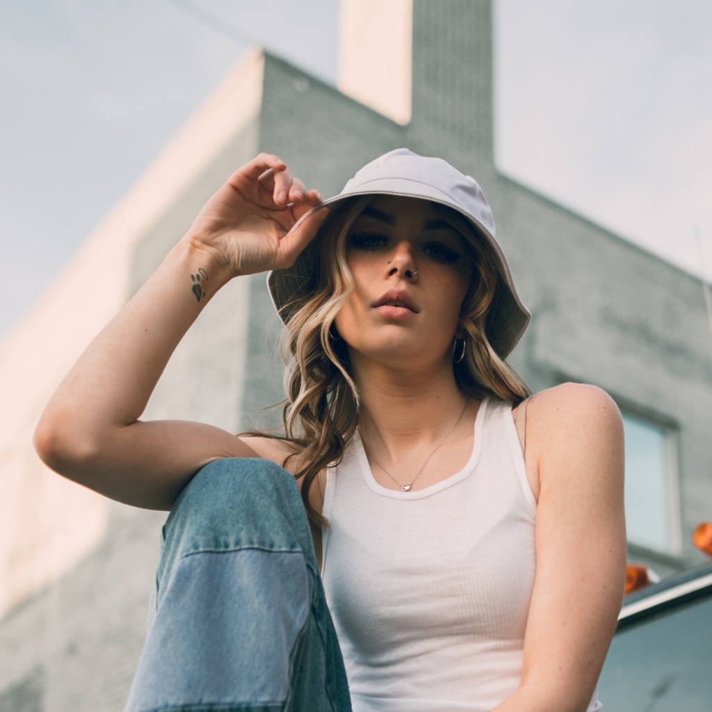 Eine stilvolle Frau mit weißem Bucket Hat, weißem Tanktop und hellblauen Jeans posiert im Freien.