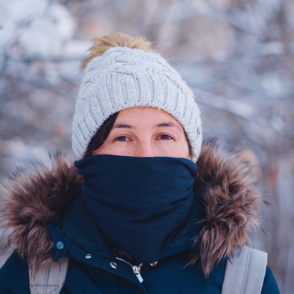 Eine Person, die eine hellgraue Strickmütze mit einem flauschigen Bommel, einen dunkelblauen balaclava-ähnlichen Schal, der Nase und Mund bedeckt, und eine marineblaue Winterjacke mit pelzbesetzter Kapuze trägt; verschneiter, verschwommener Hintergrund.<br>