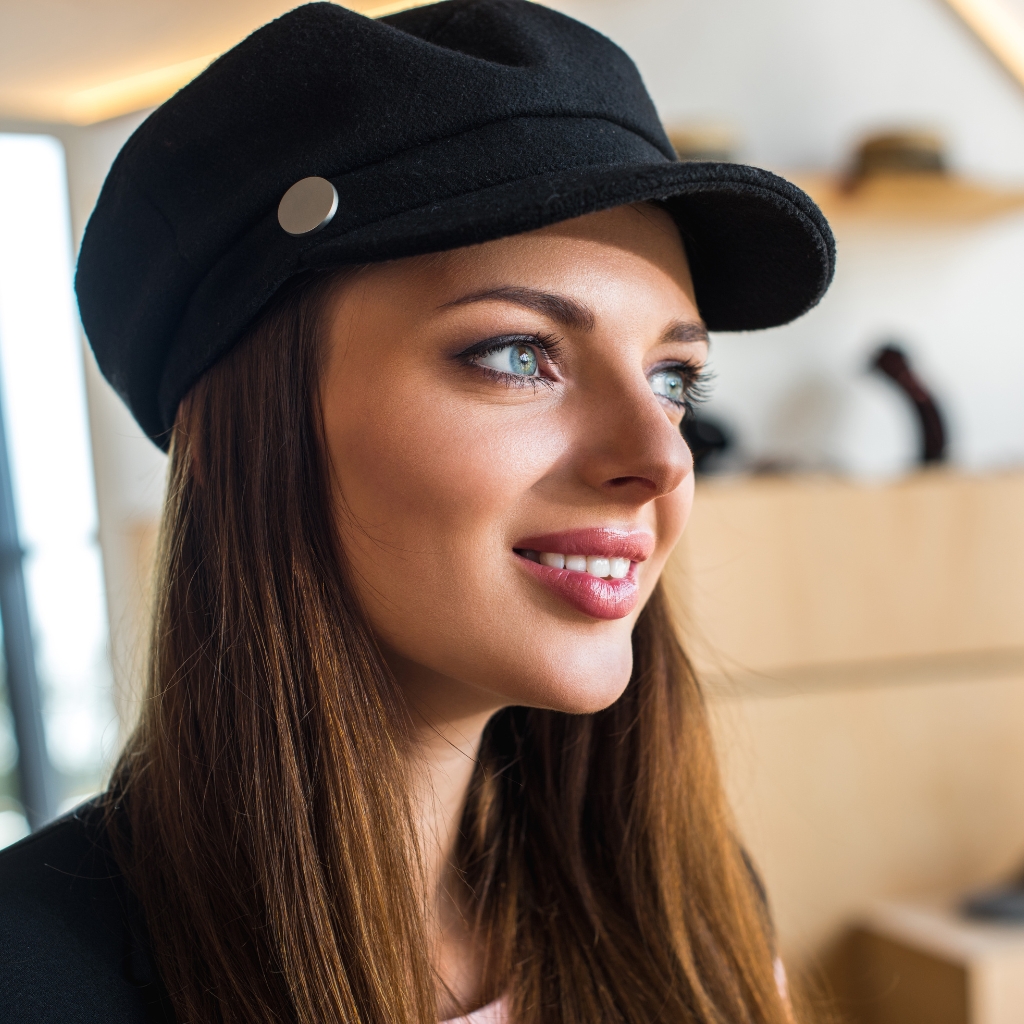 Portrait en gros plan d'une femme souriante portant une casquette en laine noire avec un bouton métallique sur le côté. Elle a de longs cheveux bruns et des yeux bleus brillants, et se tient à l'intérieur avec un arrière-plan légèrement flou. 