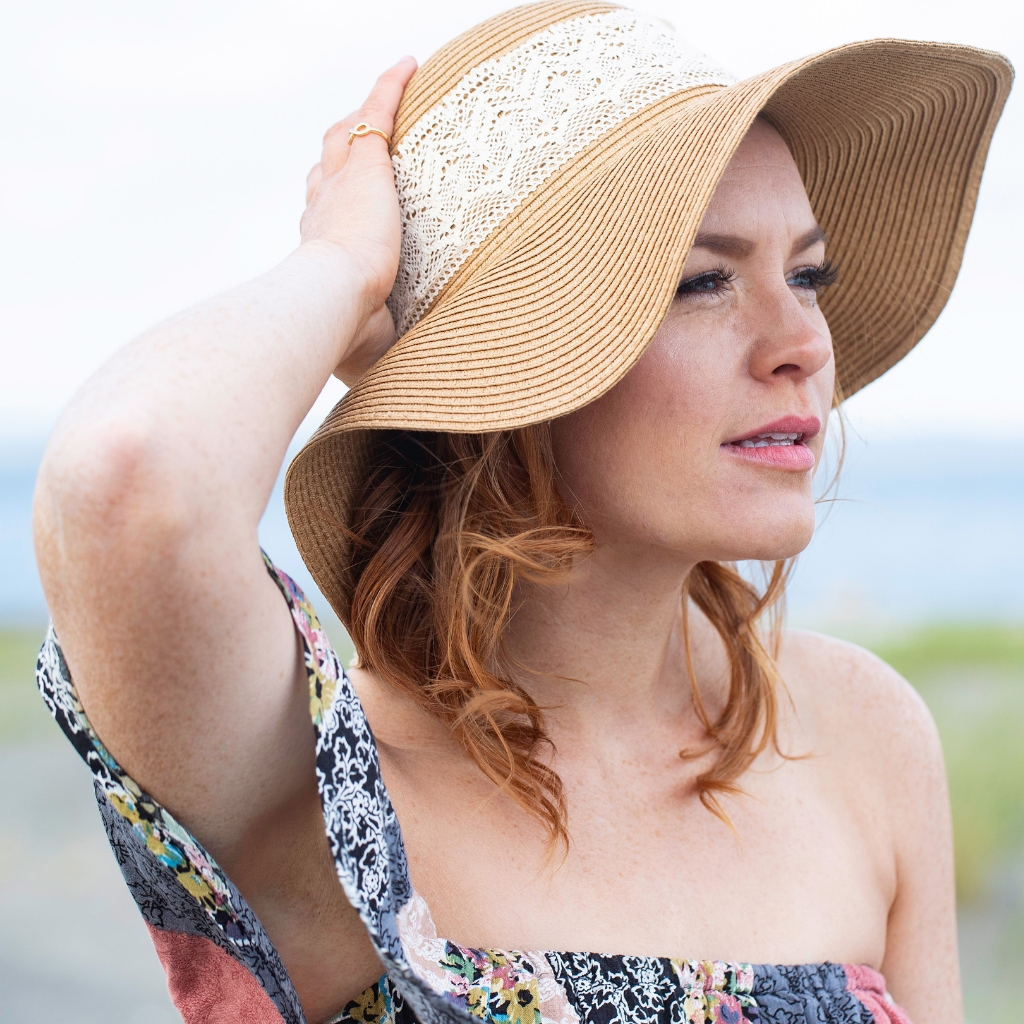 A woman at the beach wearing a wide-brim straw hat with lace detailing, gazing into the distance. She has light red hair, a floral-patterned off-shoulder dress, and a calm, sun-kissed expression.