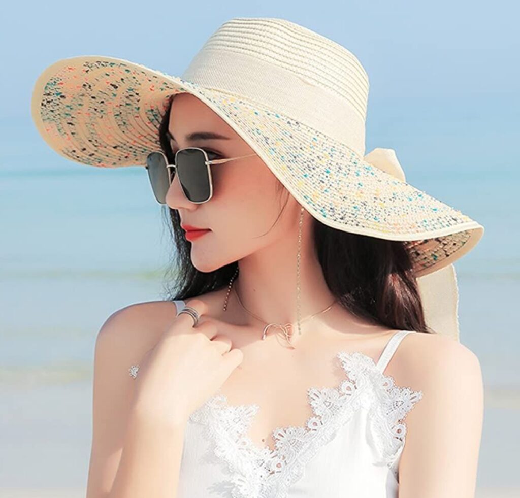 “A stylish woman wearing a wide-brim floppy straw hat, sunglasses, and a white lace-trim top at the beach, showcasing a fashionable sun hat look.”