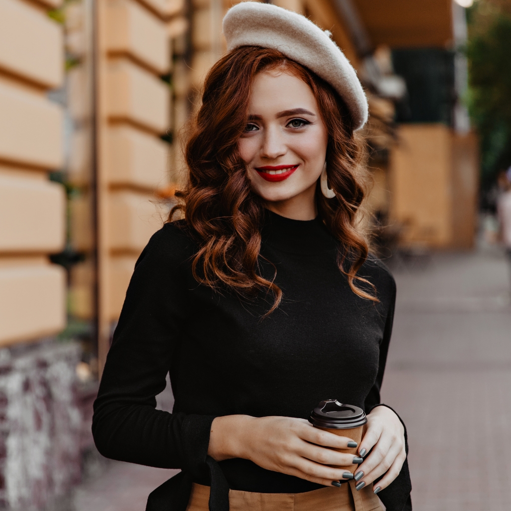 Une jeune femme élégante aux cheveux auburn ondulés, coiffée d'un béret en laine blanc et vêtue d'un col roulé noir, tenant un gobelet de café à emporter dans une rue de la ville.