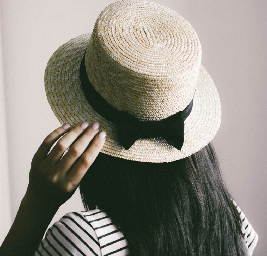 Close-up of a person wearing a woven Panama hat with a black ribbon bow, viewed from behind. The hat is light beige with a rounded crown and wide brim, and the wearer has long dark hair and a striped shirt.