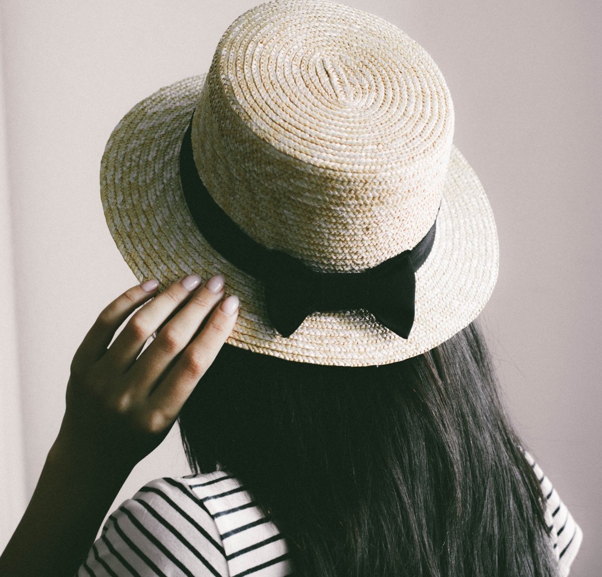 Close-up of a person wearing a woven Panama hat with a black ribbon bow, viewed from behind. The hat is light beige with a rounded crown and wide brim, and the wearer has long dark hair and a striped shirt.