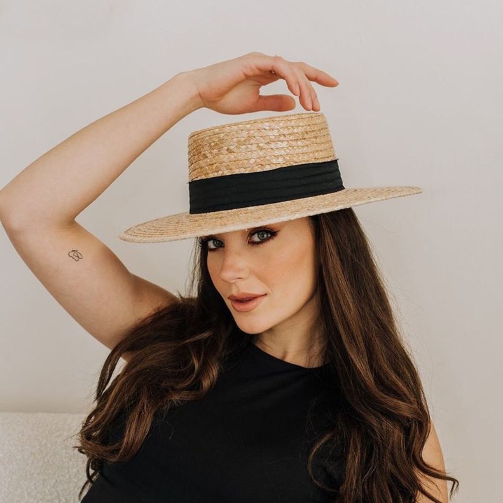 "Woman wearing a straw boater hat with a black band, posing with her hand lifting the brim."


