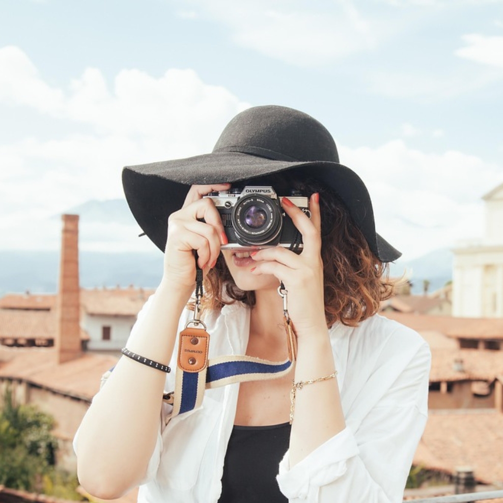 A person wearing a wide-brim black capeline hat and white shirt, holding a vintage camera up to their face as they take a photo outdoors. The background shows a sunny terrace with brick rooftops.