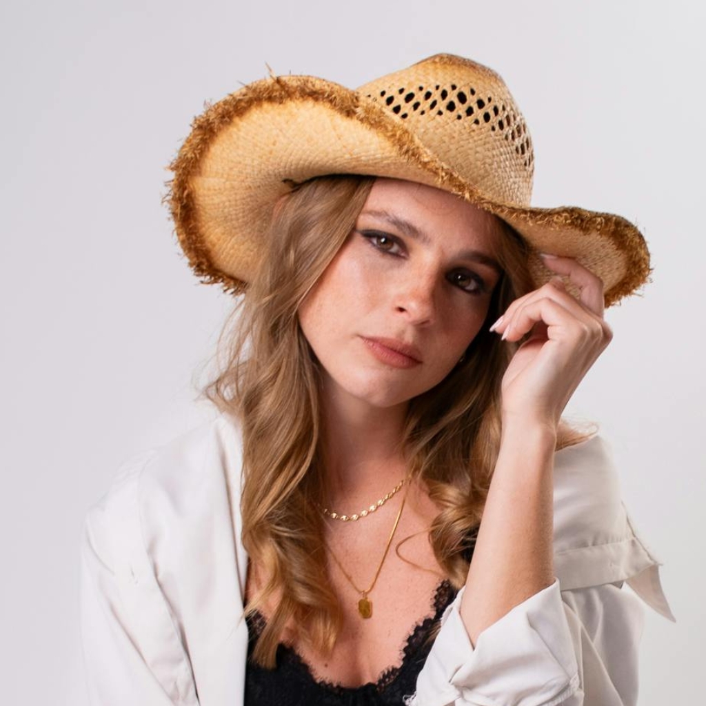 A woman wearing a woven straw cowboy beach hat, tilting her head slightly and gazing at the camera. She has wavy blonde hair, a white shirt over a black lace top, and layered gold necklaces.