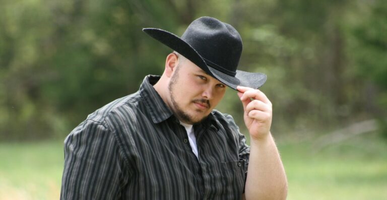 Ein Mann mit schwarzem Cowboyhut und dunkel gestreiftem Hemd, der seinen Hut lüftet, während er in einer Außenkulisse in die Kamera blickt.