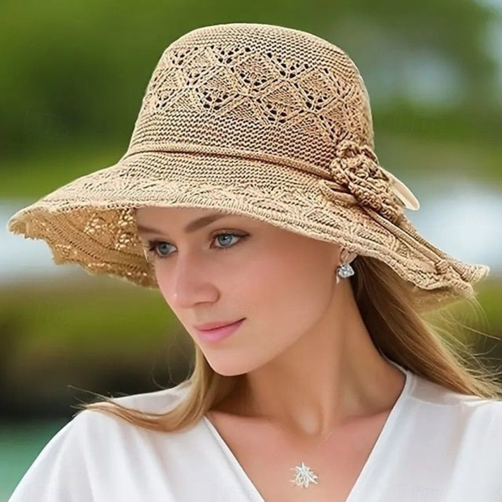 A woman wearing a wide-brim crochet beach hat in natural beige, with intricate lace detailing. She has light blue eyes, subtle jewelry (earrings and a starburst pendant), and a white top, outdoors with blurred greenery in the background.