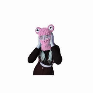 A person wearing a chunky pink knitted balaclava with two round frog-like eyes on top; the balaclava covers their head and neck, leaving the eyes visible, while the person’s hands touch the sides of the face.