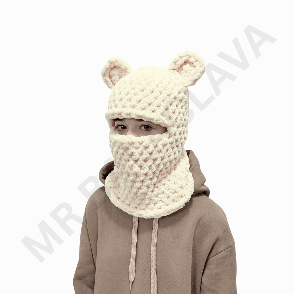 Cream-colored knit balaclava with two rounded ear-like bumps and a face opening, worn by a person in a beige hoodie.