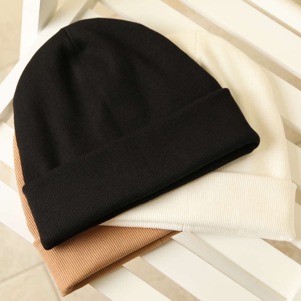 A stack of three ribbed knit beanies in black, white, and camel colors with turned-up cuffs, arranged on a white slatted surface.