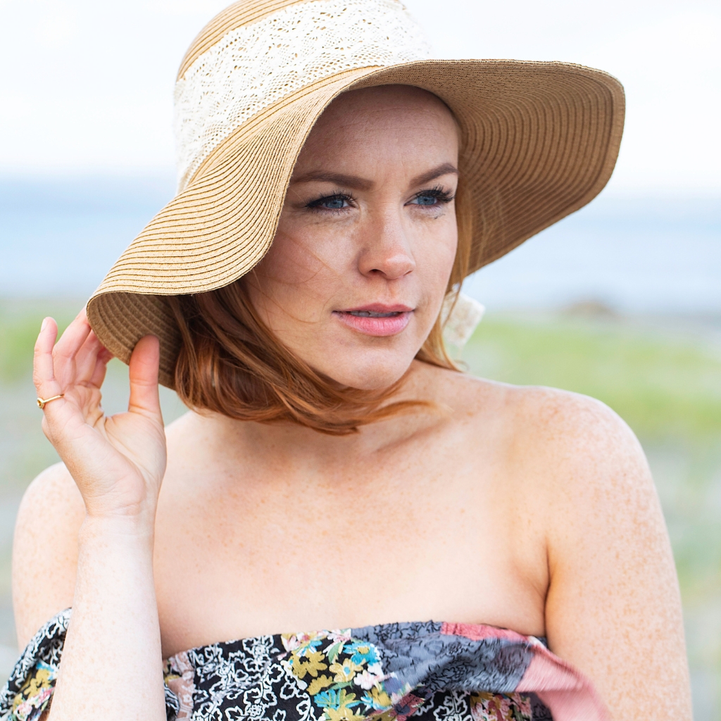A woman wearing a wide-brimmed straw floppy hat outdoors, with light brown hair, light skin with freckles, and an off-shoulder floral dress, touching the hat brim.


