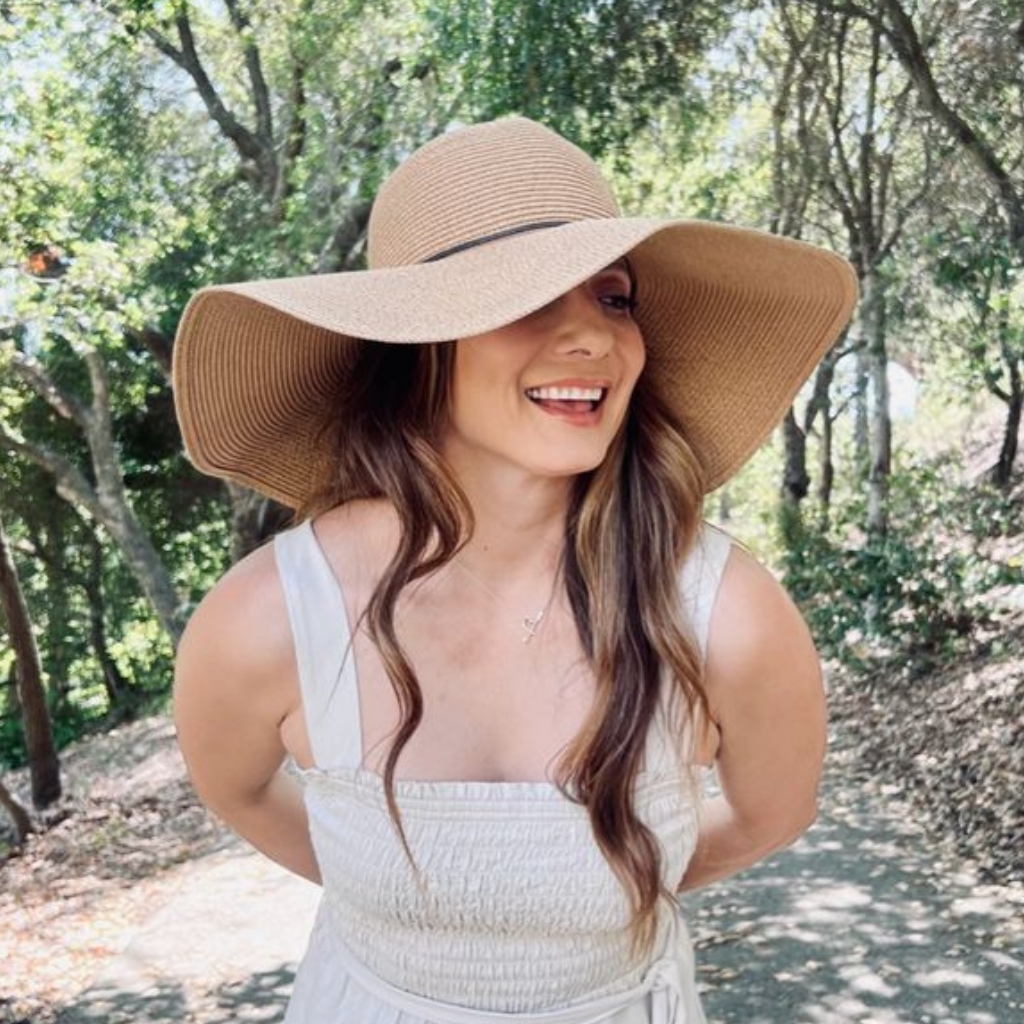 Une femme souriante portant un chapeau de paille à large bord, une robe sans manches de couleur claire, à l'extérieur parmi les arbres par une journée ensoleillée.