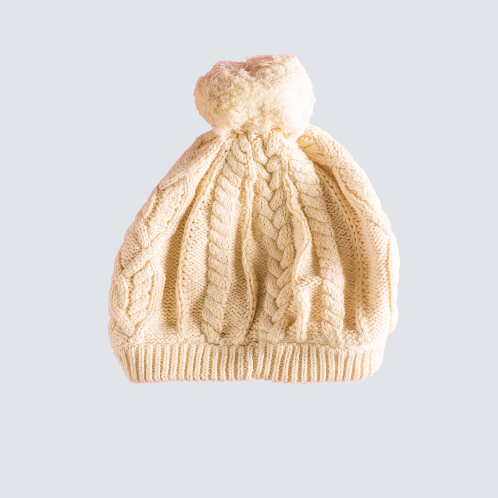 Cream-colored knitted beanie with a large pom-pom on top, featuring a cable-knit pattern and ribbed brim, set against a light gray background.