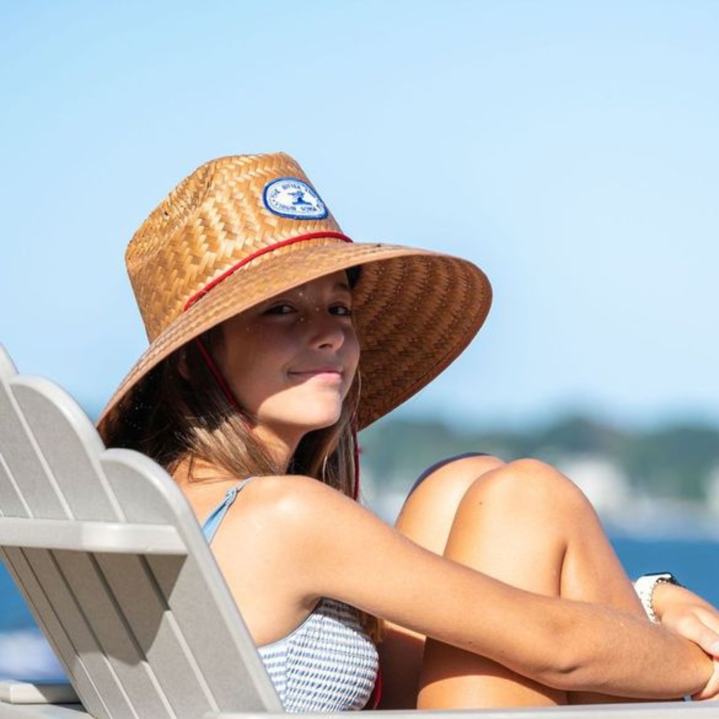 A young lifeguard seated on a lounge chair at the beach, wearing a wide-brimmed straw hat and a light blue checkered top, with sunlit skin and a calm expression.