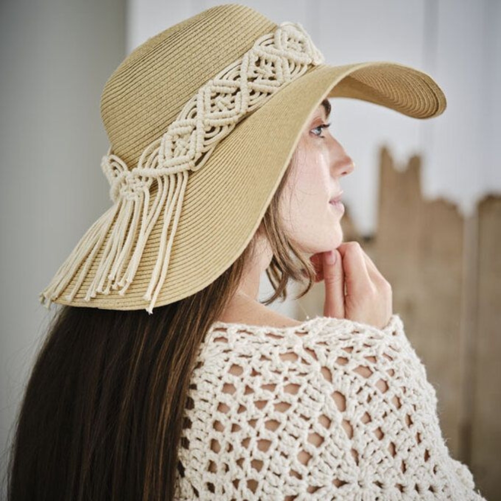 A woman wearing a wide-brimmed straw hat with decorative macramé cord detailing around the crown. She has long dark hair and wears a cream crochet top, posing in profile.