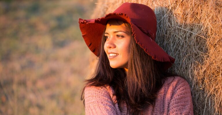 Eine lächelnde Frau mit langen braunen Haaren, die einen roten Hut mit breiter Krempe und einen rosa Pullover trägt und sich während der goldenen Stunde im Freien an einen Heuballen lehnt.