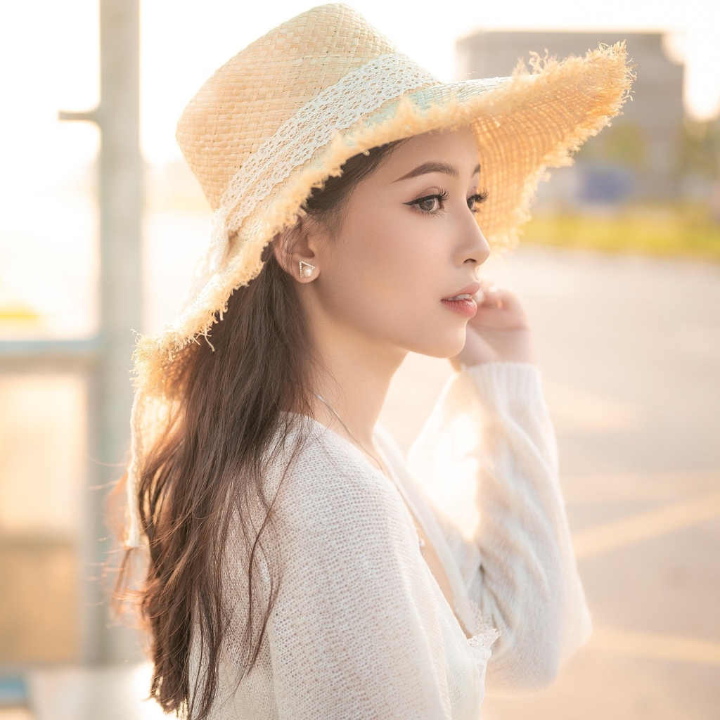 "Profile of a young woman wearing a wide straw hat with a frayed brim, light-colored sweater, and subtle makeup in soft warm sunlight."

