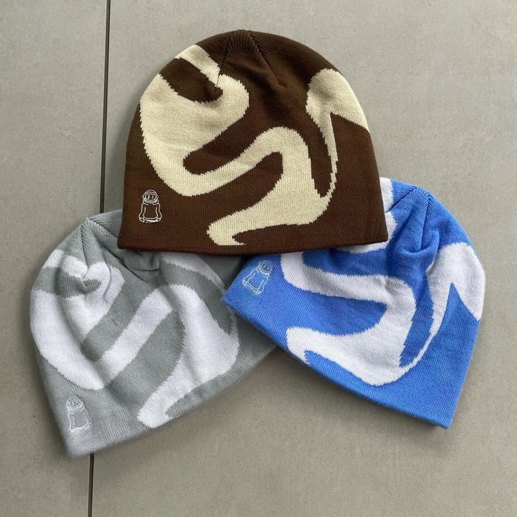 Three knit beanies arranged on a neutral surface: one brown with a cream abstract pattern on top, a light gray beanie with a white pattern to the left, and a blue beanie with a white pattern on the right; each features a small embroidered astronaut badge near the edge. Custom Graphic Beanie Manufacturer